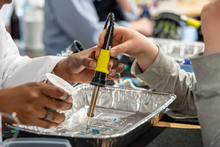 Nahaufnahme einer Kinderhand und einer erwachsenen Hand, die gemeinsam mit einem Lötkolben an kleinen elektronischen Bauteilen arbeiten; der Lötkolben wird geführt, während Bauteile in einer Aluminiumschale positioniert sind. Im Hintergrund ist unscharf eine Lernumgebung zu erkennen.
