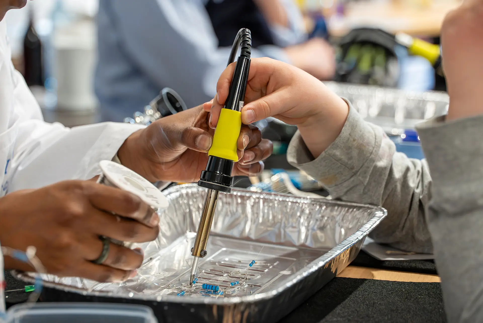 Nahaufnahme einer Kinderhand und einer erwachsenen Hand, die gemeinsam mit einem Lötkolben an kleinen elektronischen Bauteilen arbeiten; der Lötkolben wird geführt, während Bauteile in einer Aluminiumschale positioniert sind. Im Hintergrund ist unscharf eine Lernumgebung zu erkennen.